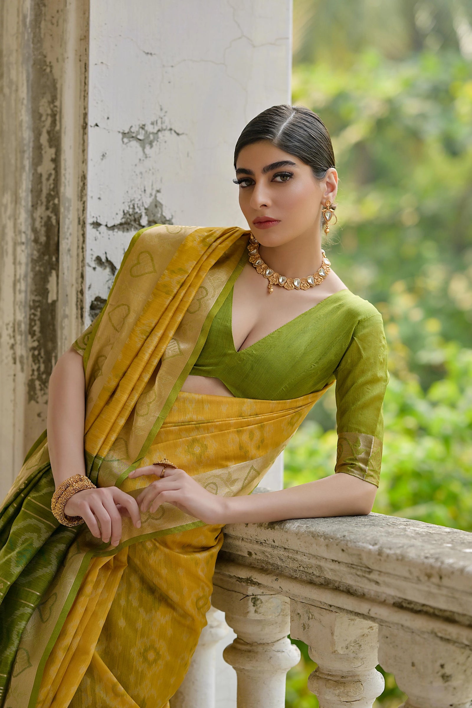 Woman in a green and yellow saree leaning against a stone wall with greenery in the background