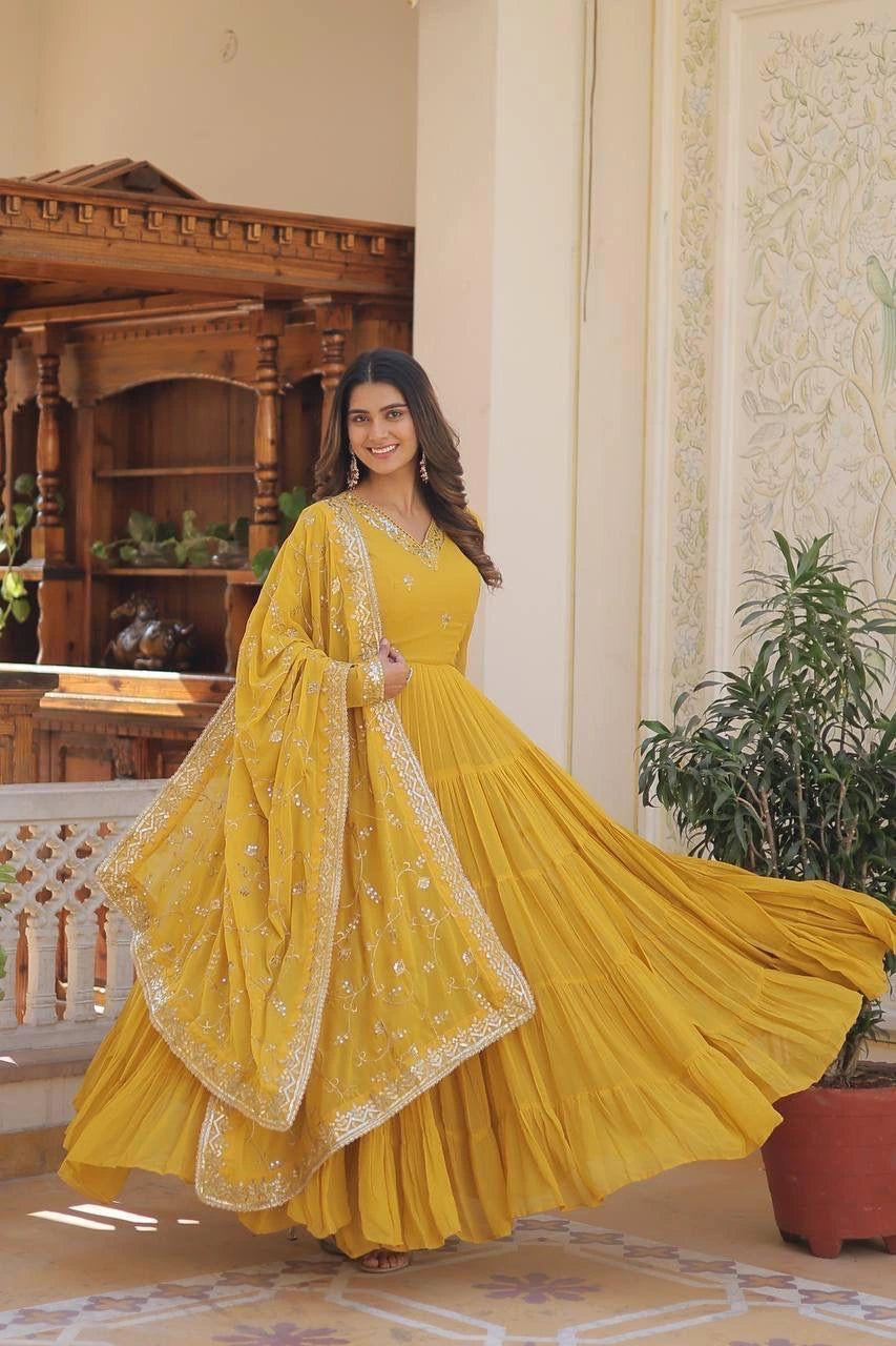 Woman in a yellow traditional outfit standing in an elegant room with wooden furniture and decorative plants.