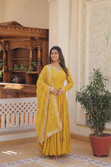 Woman in a yellow traditional outfit standing in an ornate room with wooden furniture and decorative wall.