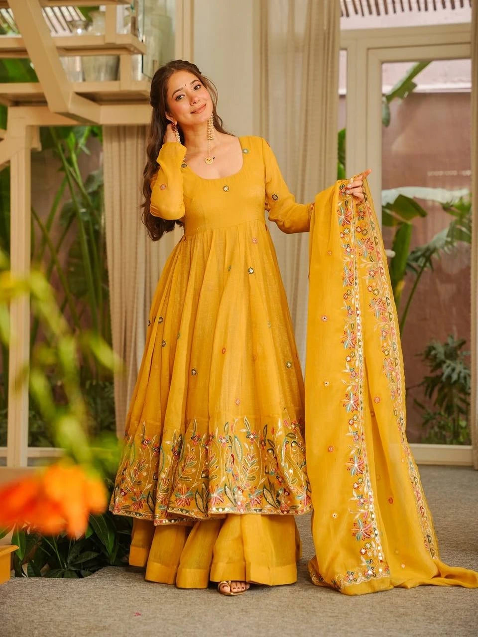 Woman in a yellow traditional outfit with a floral dupatta, standing indoors.