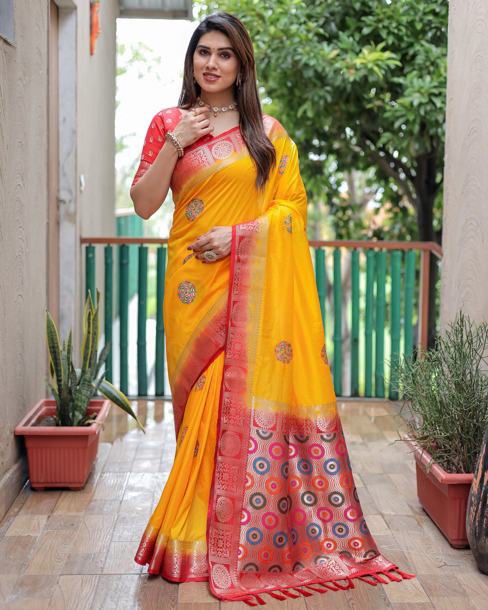 Woman wearing a yellow and red saree with a patterned border, standing on a wooden floor with plants in the background.