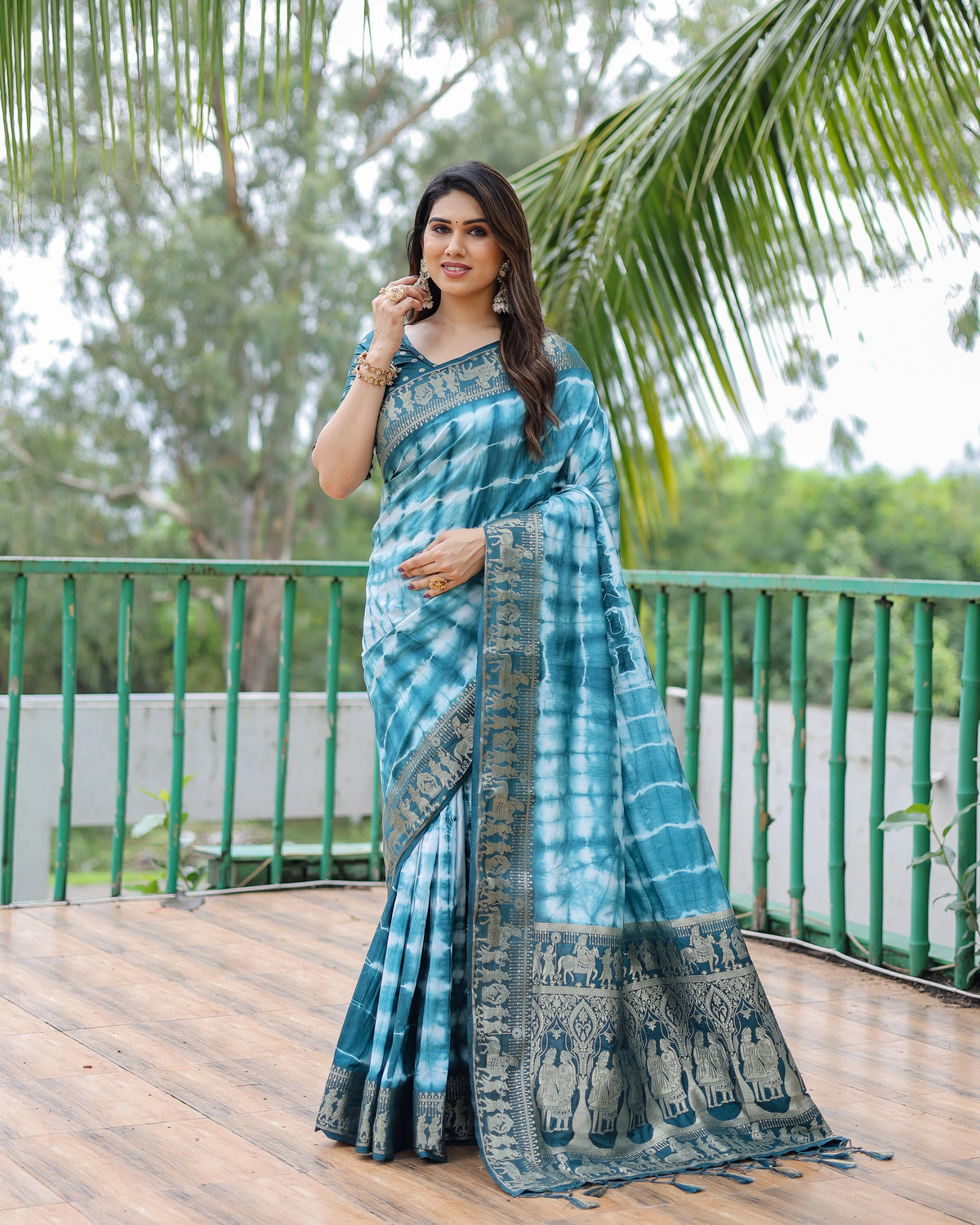 Woman wearing a blue and green saree standing outdoors with greenery in the background