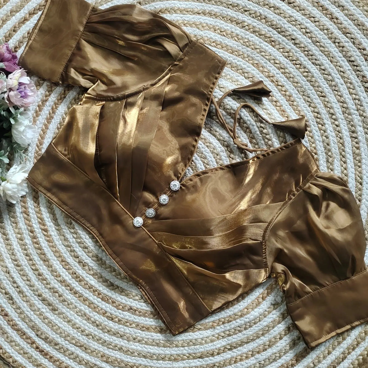 Dark Brown blouse with pleated detailing and three decorative silver buttons on the front, featuring elbow-length sleeves and tie-up tassels, displayed on a circular woven mat with white and pink flowers on the side.