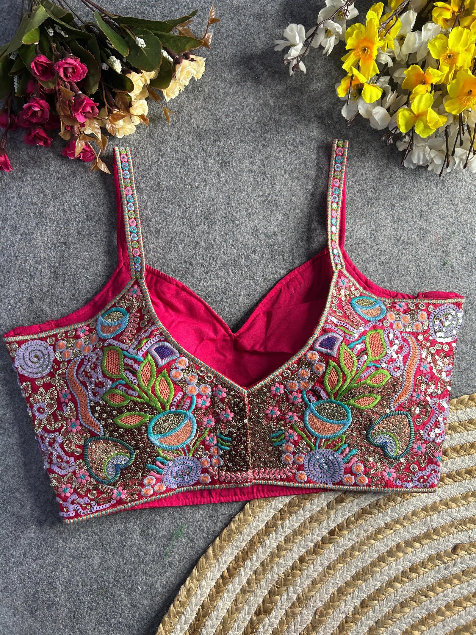 aditional multicolor bustier-style blouse featuring intricate thread embroidery and sequin embellishments in floral and paisley motifs, with structured cups and adjustable straps, displayed flat on a textured grey surface surrounded by artificial pink, yellow, and white flowers.