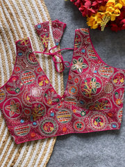  Traditional magenta blouse featuring vibrant multicolor thread embroidery and mirror work in circular floral and geometric patterns, with a deep V-neck and tie-back detail, displayed flat on a textured mat with artificial red and yellow flowers in the corner.