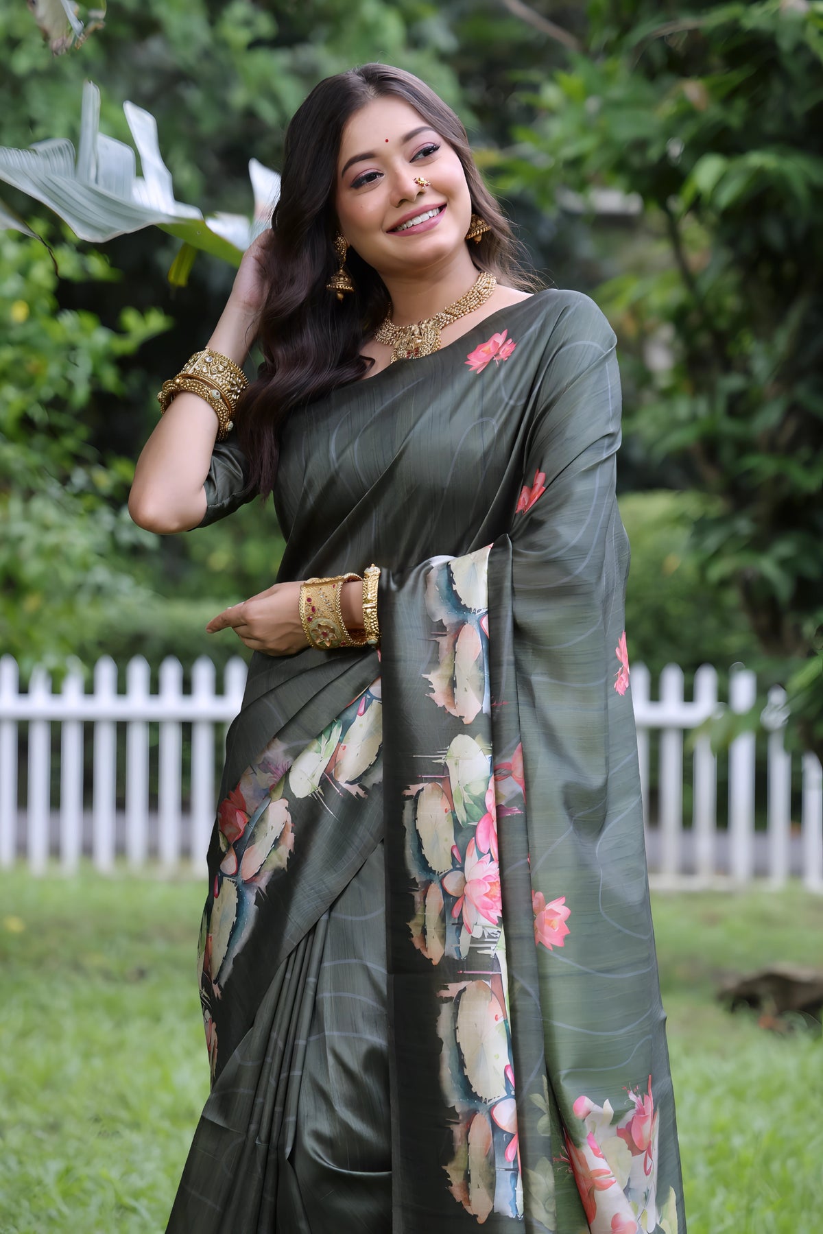 A woman models a stunning Dark Olive Green Soft Tussar Silk Saree. The saree features a large-scale digital print of colorful floral and lotus motifs in pink, white, and pale blue on the pallu and lower drape. She wears a matching elbow-sleeve green blouse and heavy gold Kundan jewelry, posing outdoors in a garden setting.
