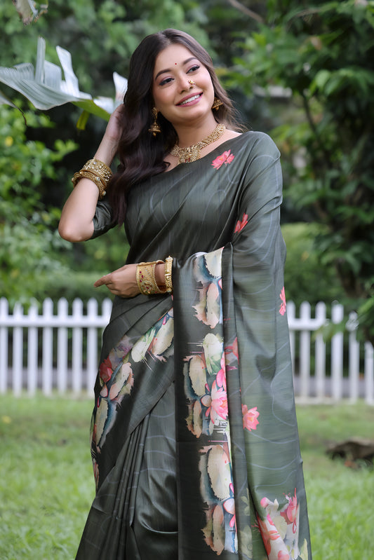 A woman models a stunning Dark Olive Green Soft Tussar Silk Saree. The saree features a large-scale digital print of colorful floral and lotus motifs in pink, white, and pale blue on the pallu and lower drape. She wears a matching elbow-sleeve green blouse and heavy gold Kundan jewelry, posing outdoors in a garden setting.