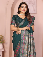 Woman wearing a rich teal green silk saree with an ornate multicolor ethnic border and pallu featuring traditional motifs in pink, silver, and green. The saree is paired with a matching short-sleeve blouse adorned with intricate embellishments. She accessorizes with a statement choker necklace, bangles, and rings, posing elegantly in a pastel arched studio with potted green plants, minimalist decor, and a white side table.