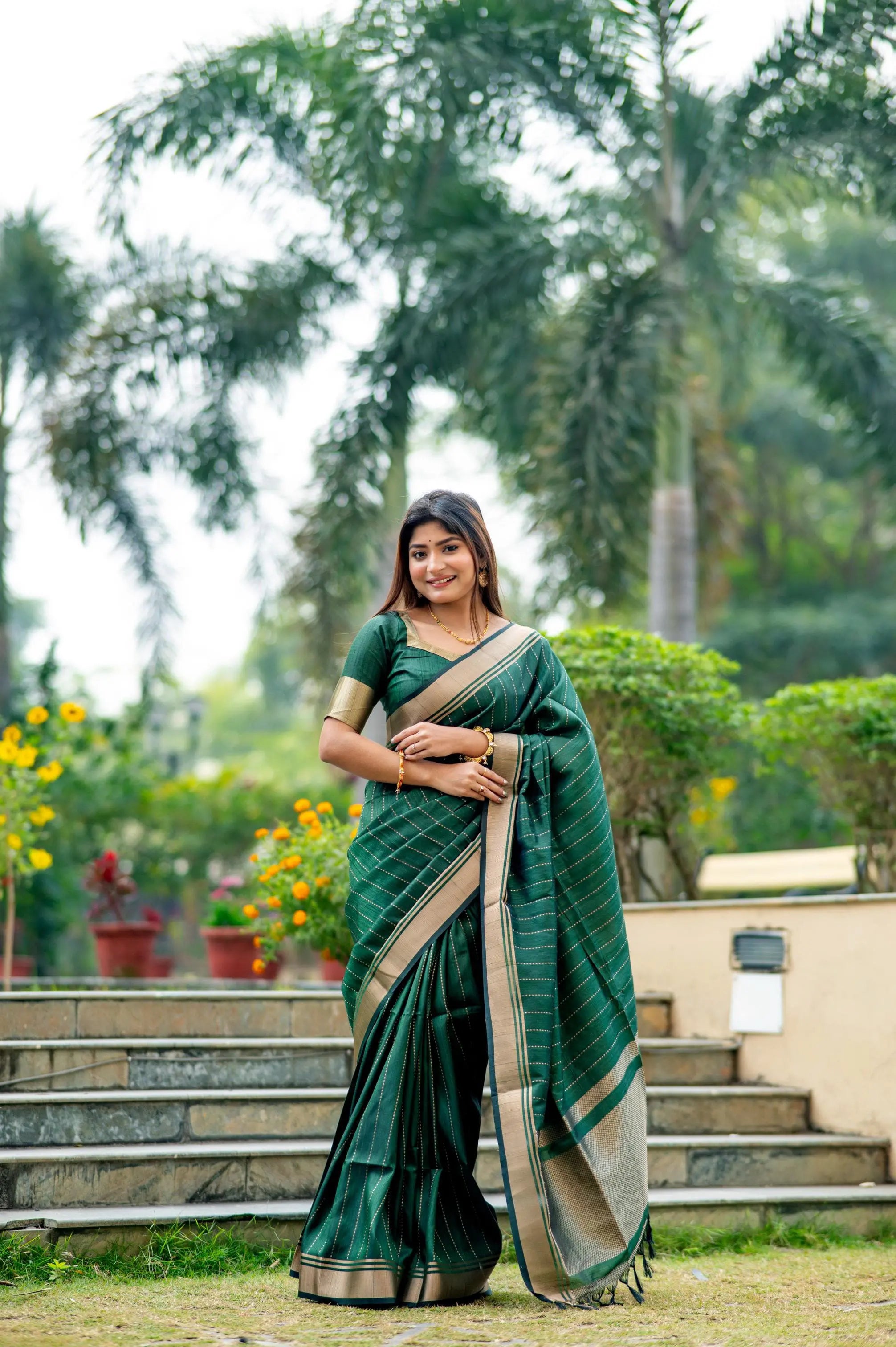 Woman in a dark green saree with zari work standing outdoors on stone steps with greenery in the background