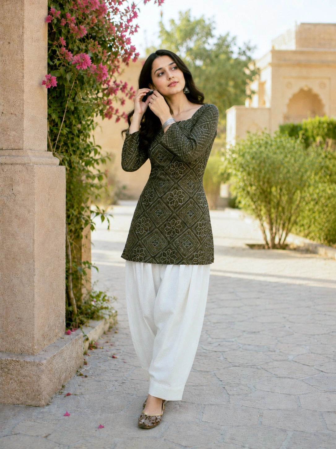 Woman in a green patterned top and white pants standing outdoors near a building with greenery.