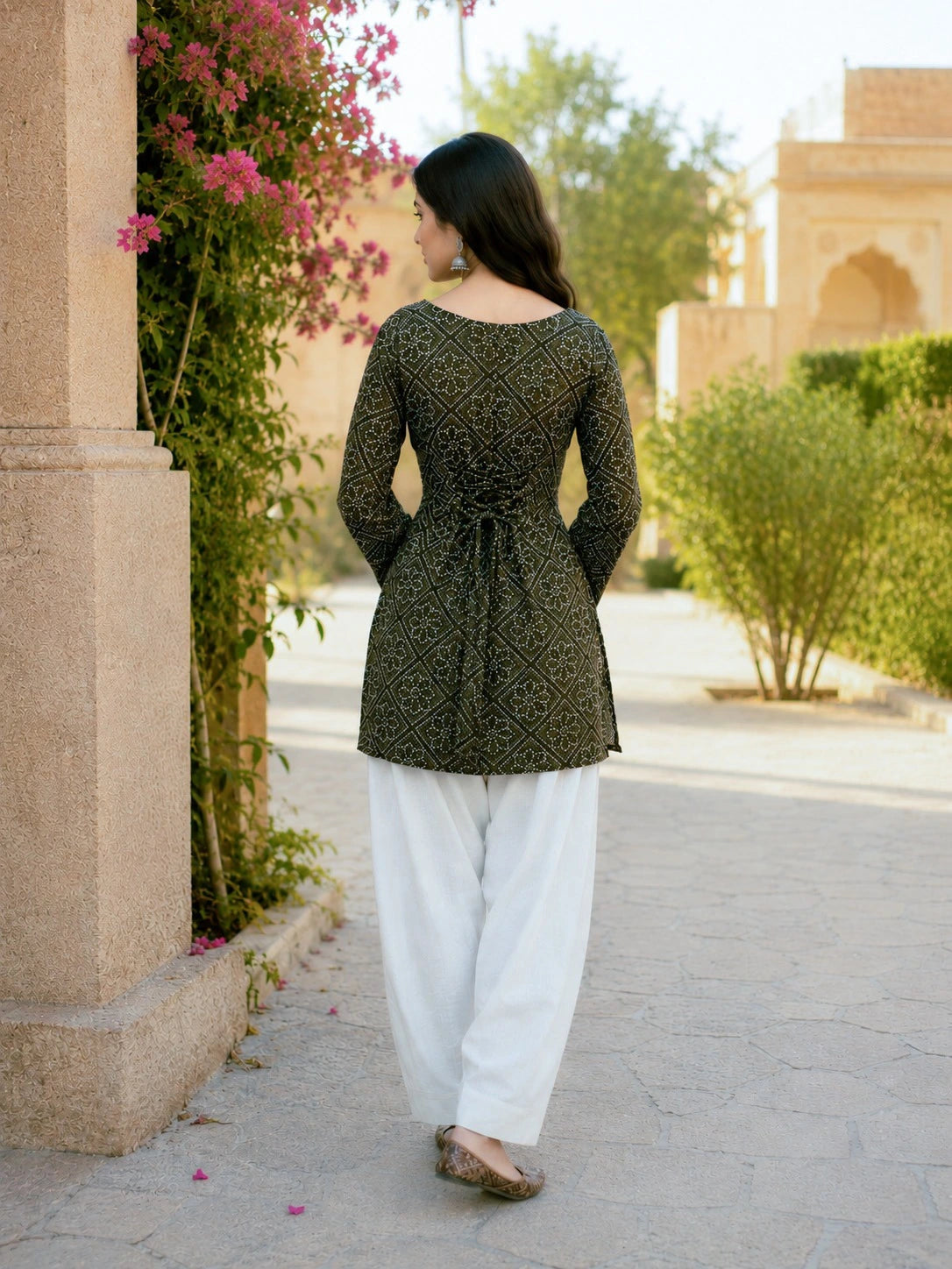 Woman in a green patterned top and white pants walking outdoors near a building with plants.