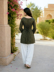 Woman in a green patterned top and white pants walking outdoors near a building with plants.