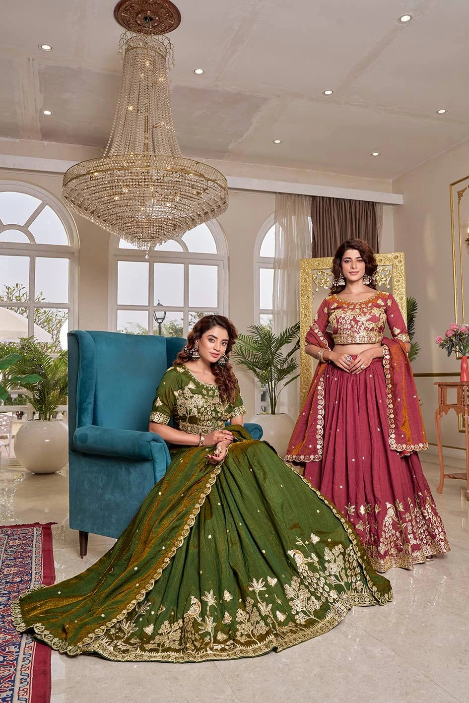 Two women in traditional green and pink lehengas sitting in a stylish room with a chandelier.