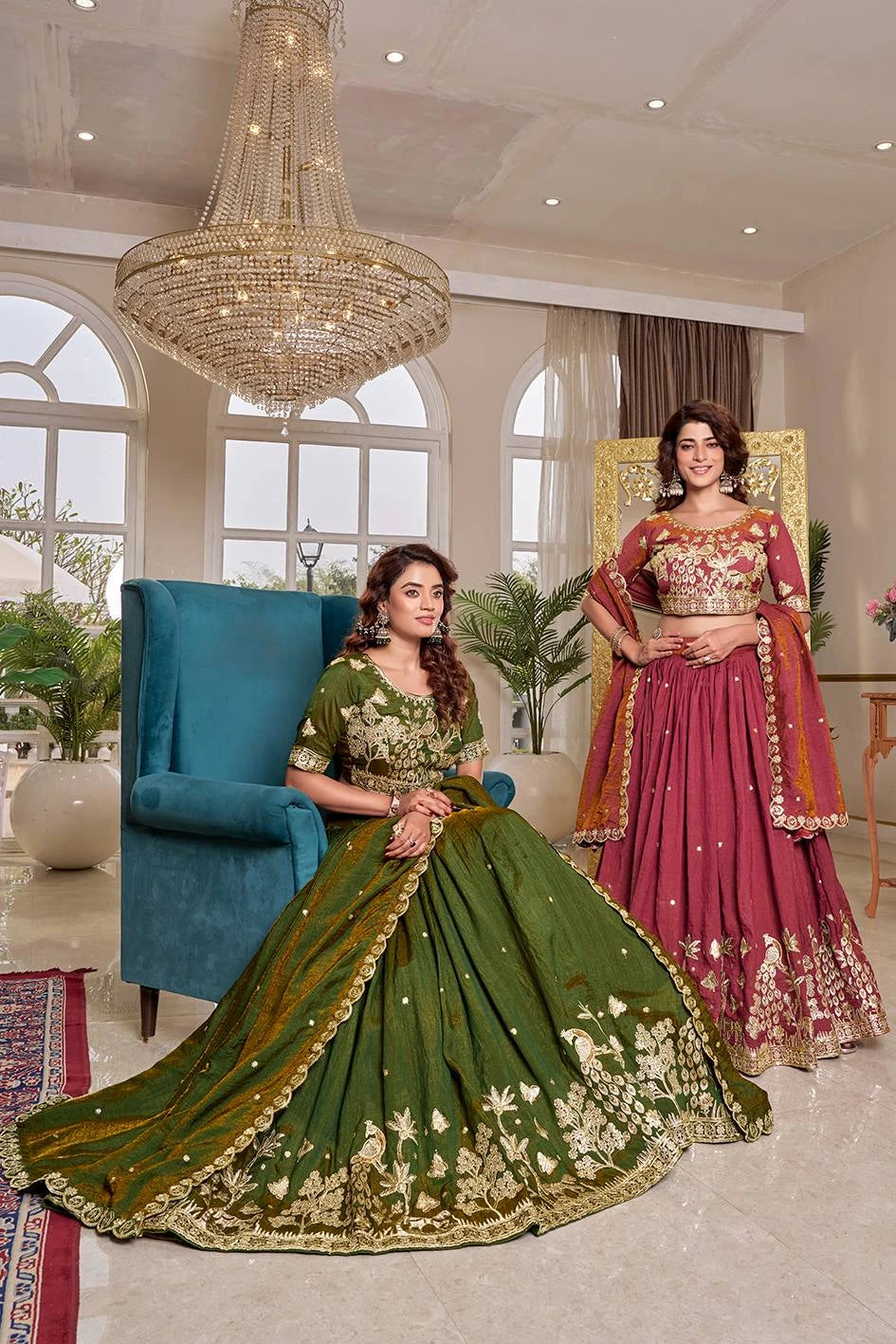 Two women in traditional green and pink lehengas sitting in a well-lit room with a chandelier.