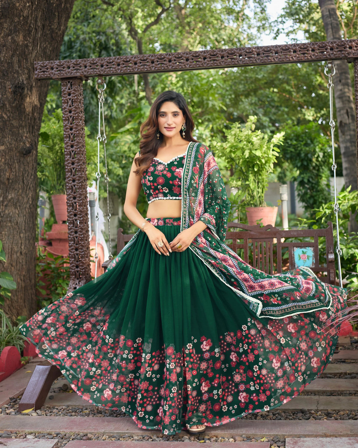 Woman in a green and pink floral lehenga standing outdoors with a swing and trees in the background.
