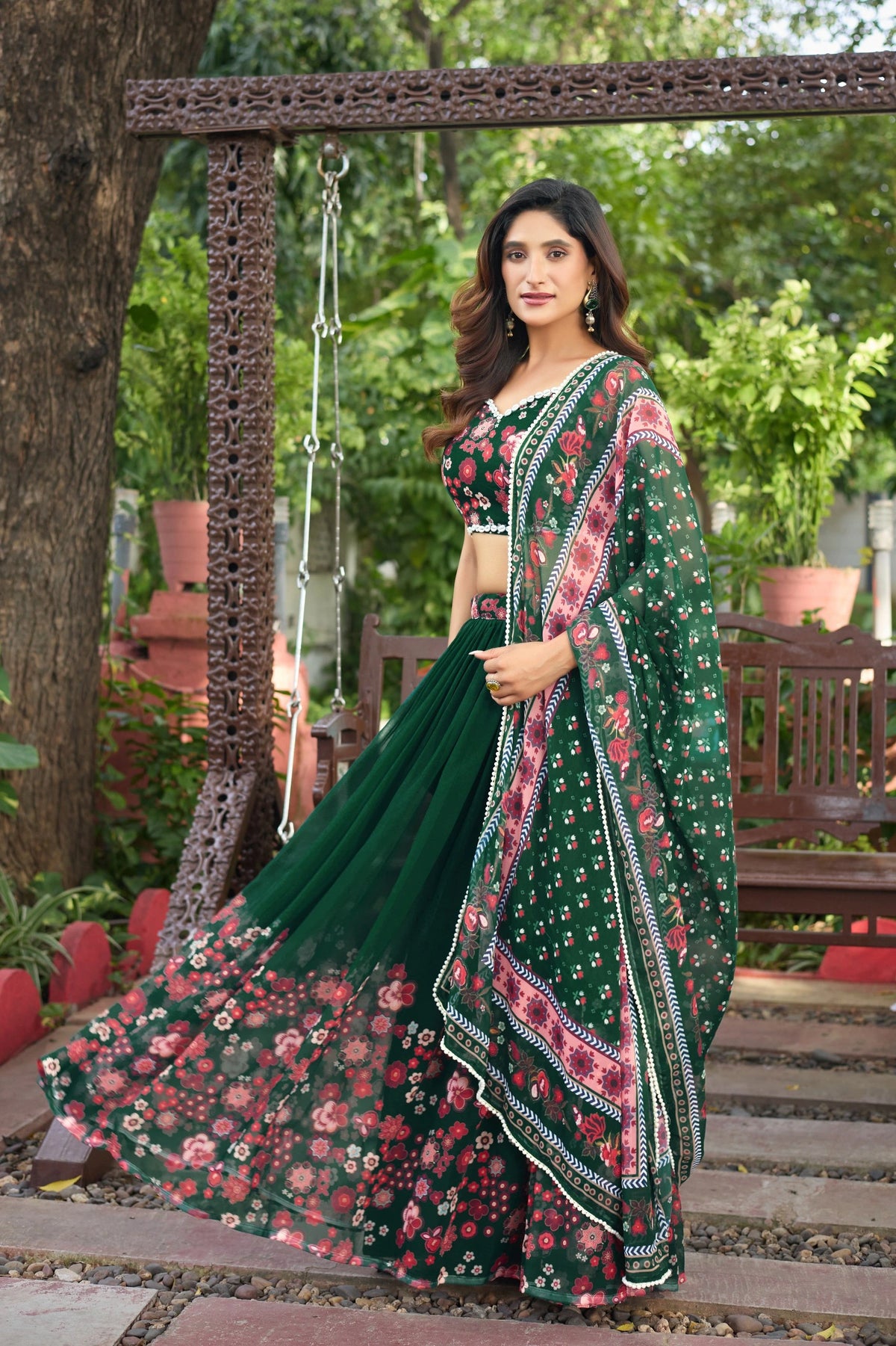 Woman in a green floral saree standing outdoors with plants and wooden furniture in the background