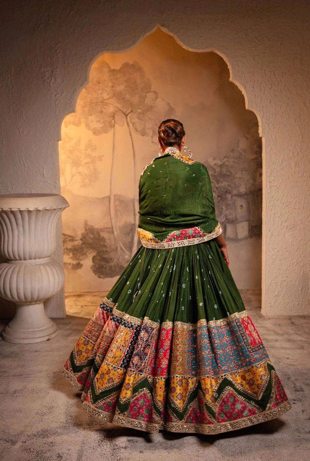 Person wearing a traditional green and multicolored dress with intricate patterns, standing in front of an ornate archway.