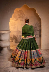 Person wearing a traditional green and multicolored dress with intricate patterns, standing in front of an ornate archway.
