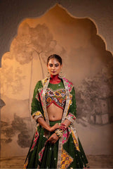 Woman in traditional green and multicolored outfit with jewelry against a decorative wall.