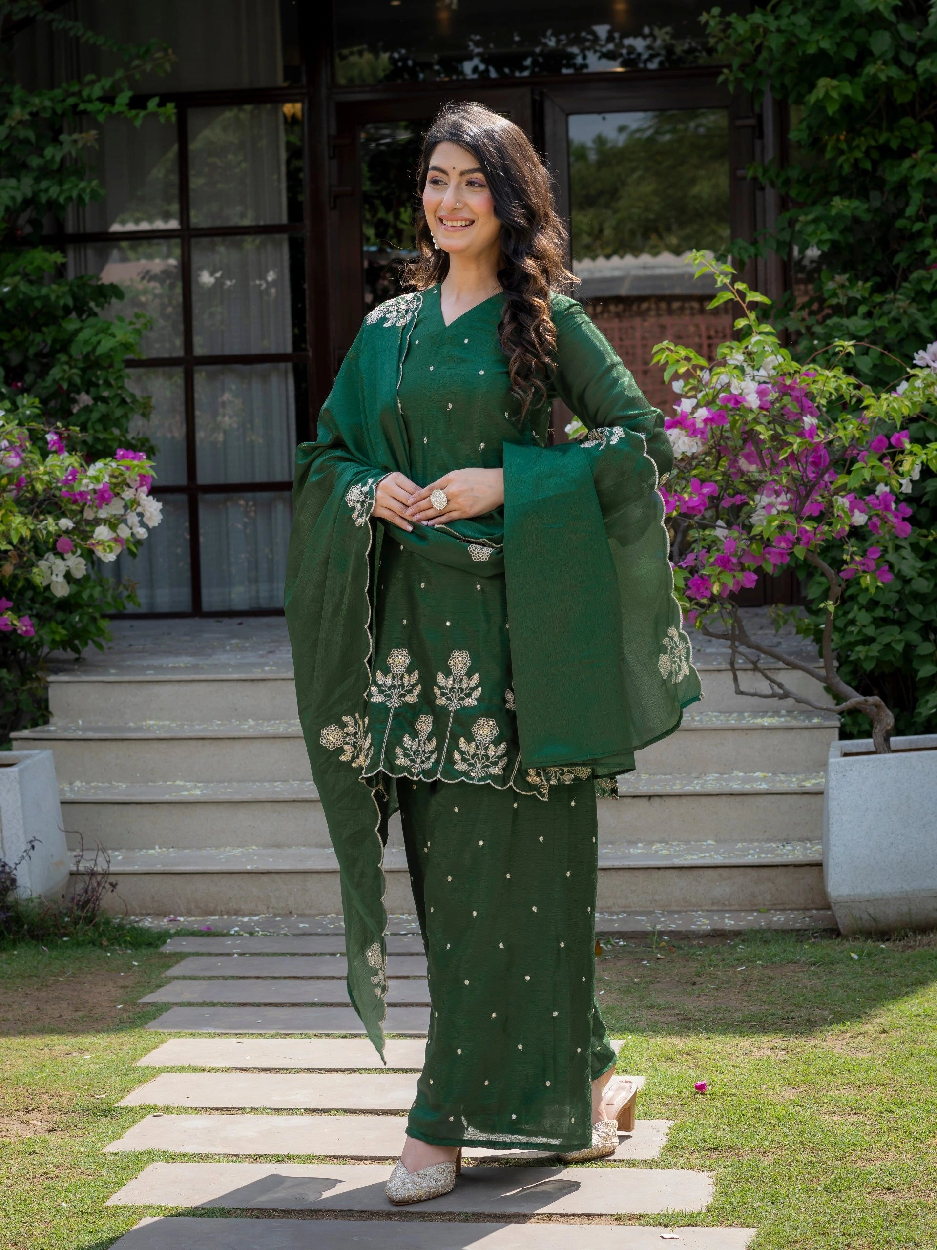 Woman in a green traditional outfit standing outdoors with flowers and steps in the background