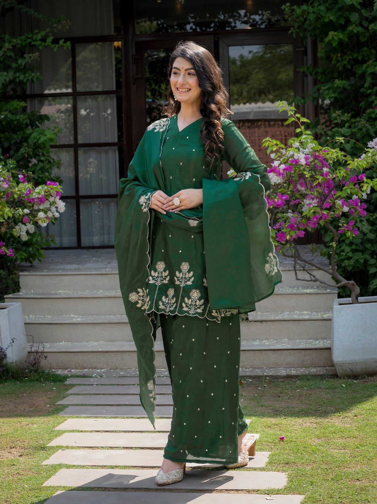 Woman in a green traditional outfit standing outdoors with flowers and steps in the background