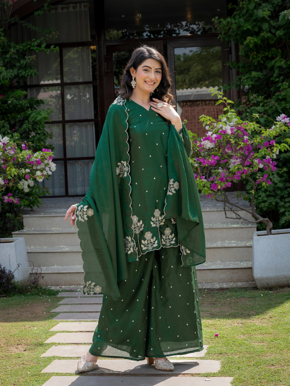 Woman in a green traditional outfit standing outdoors with flowers and a building in the background