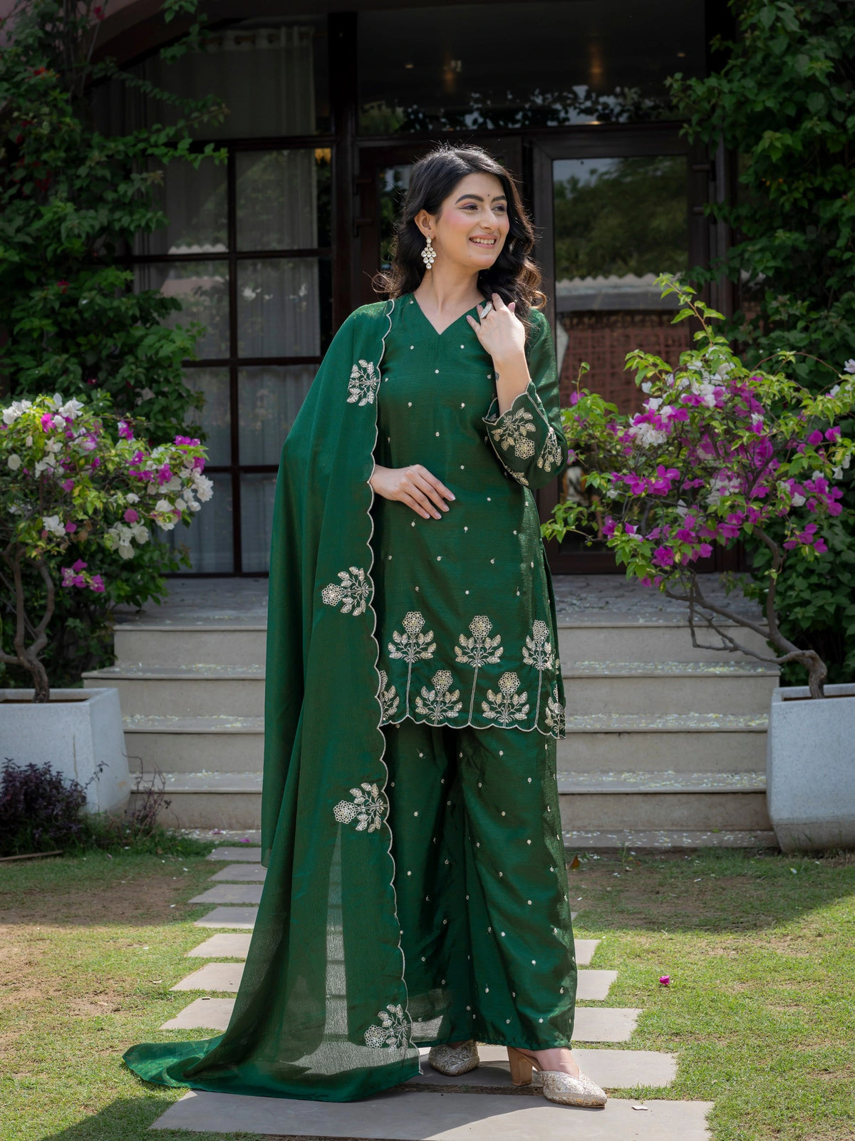Woman in a green traditional outfit standing outdoors with flowers and a building in the background
