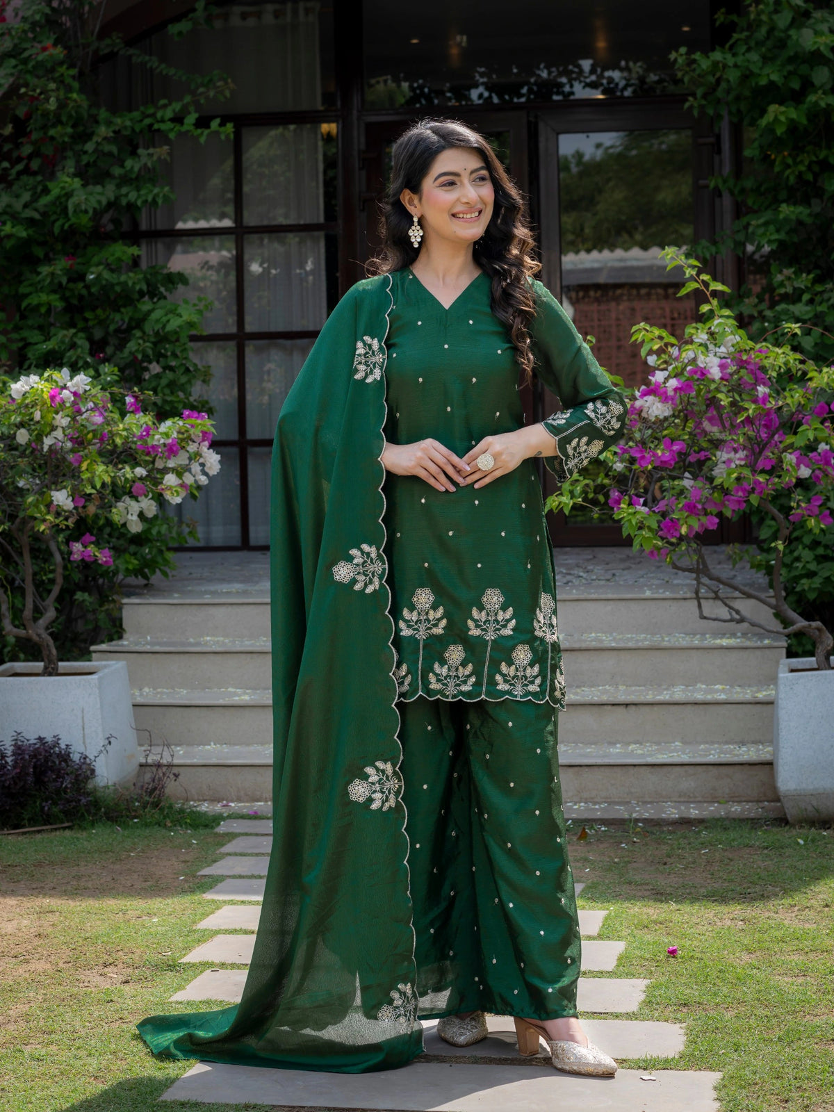 Woman in a green traditional outfit standing outdoors with flowers and steps in the background