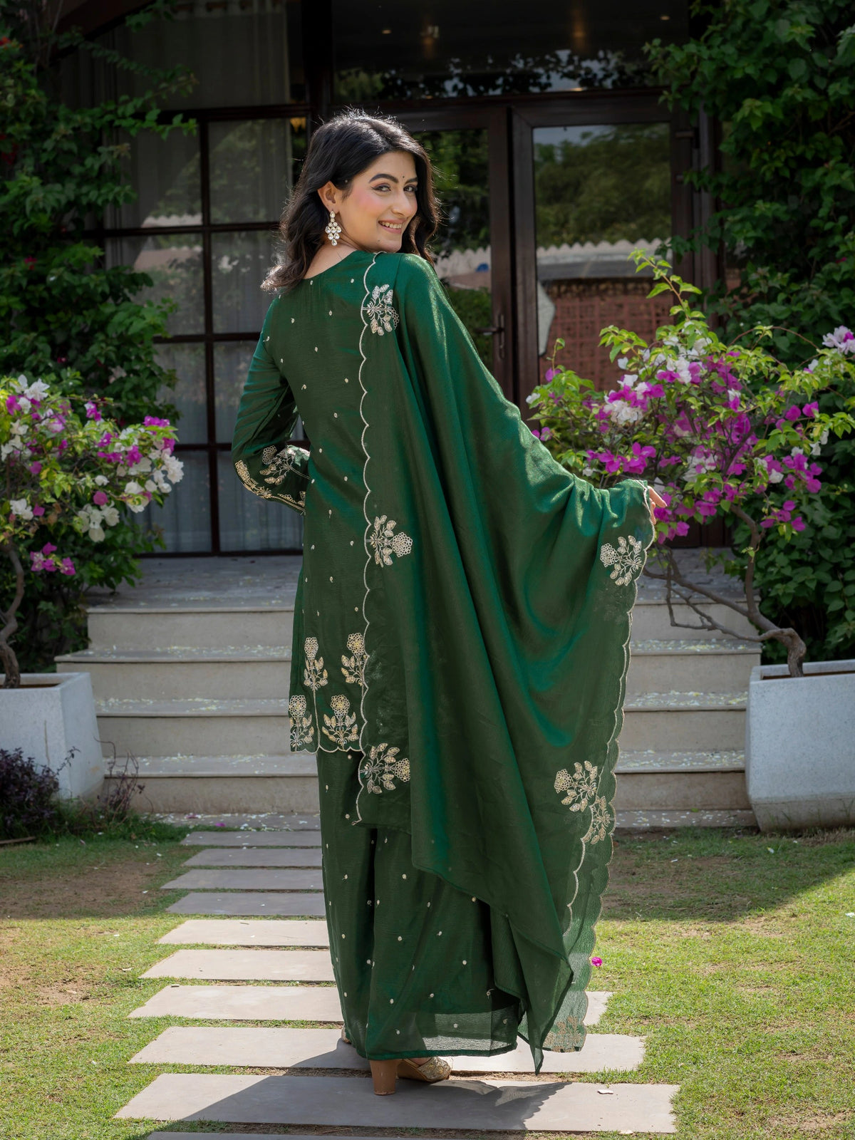 Woman in a green traditional outfit standing outdoors with flowers and a building in the background