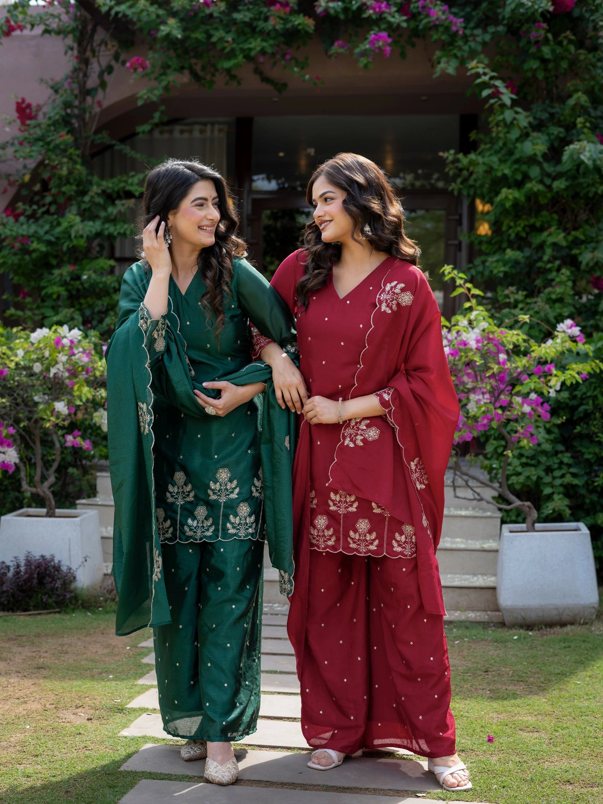 Two women in traditional green and red outfits standing outdoors with greenery and flowers in the background.