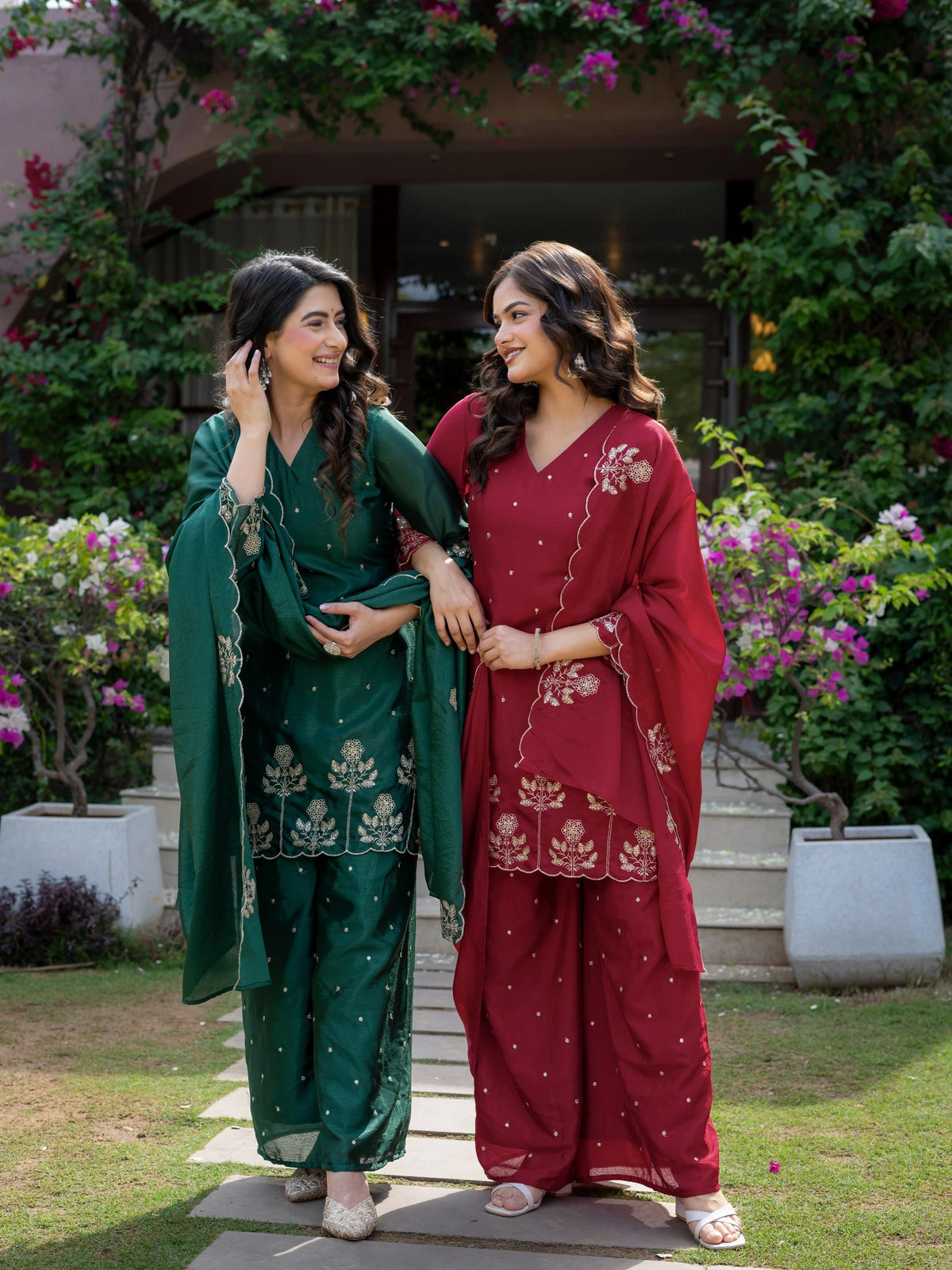 Two women in traditional green and red outfits standing outdoors with greenery and flowers in the background.
