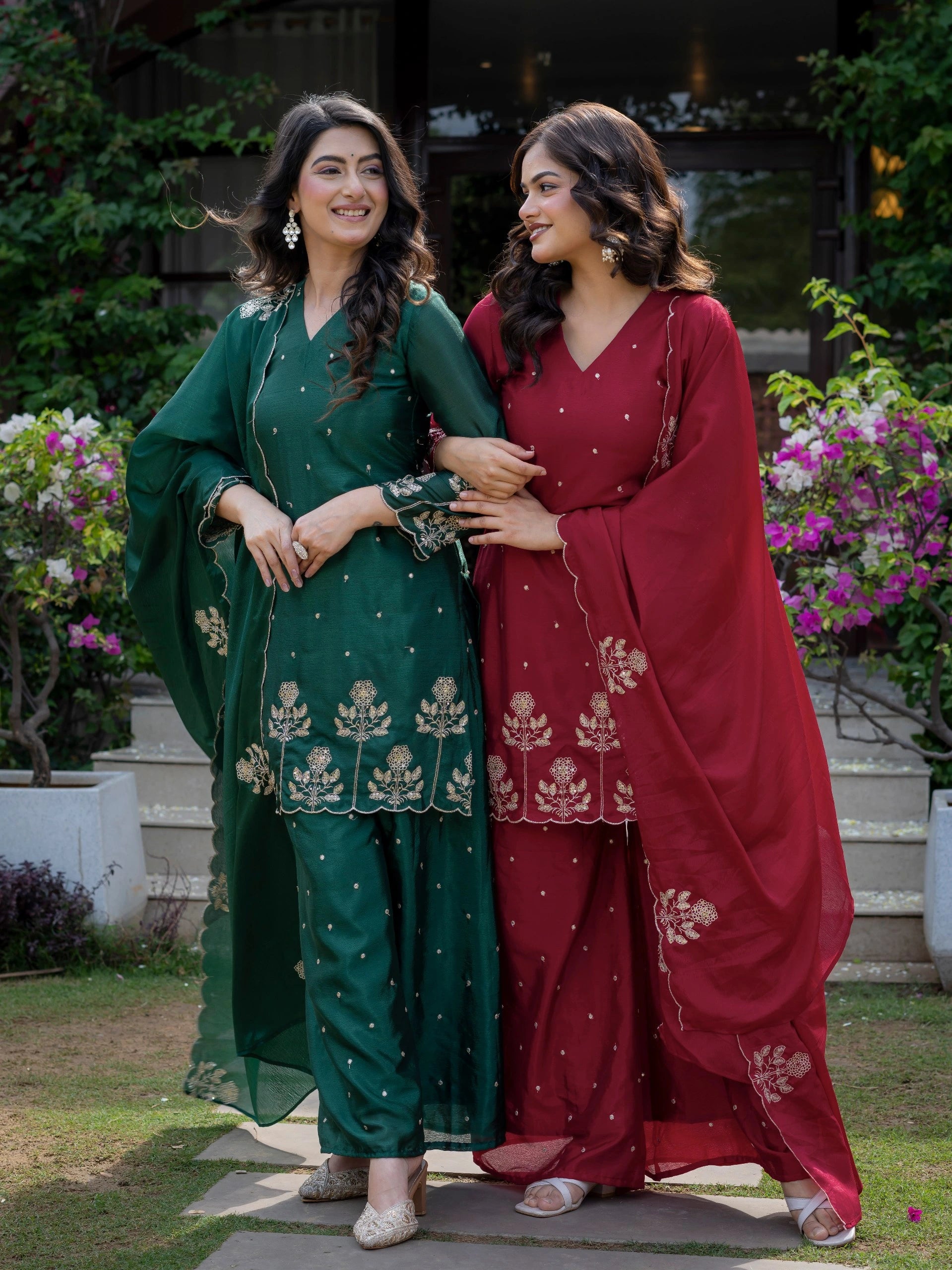 Two women in traditional green and red outfits standing outdoors with flowers and plants in the background.
