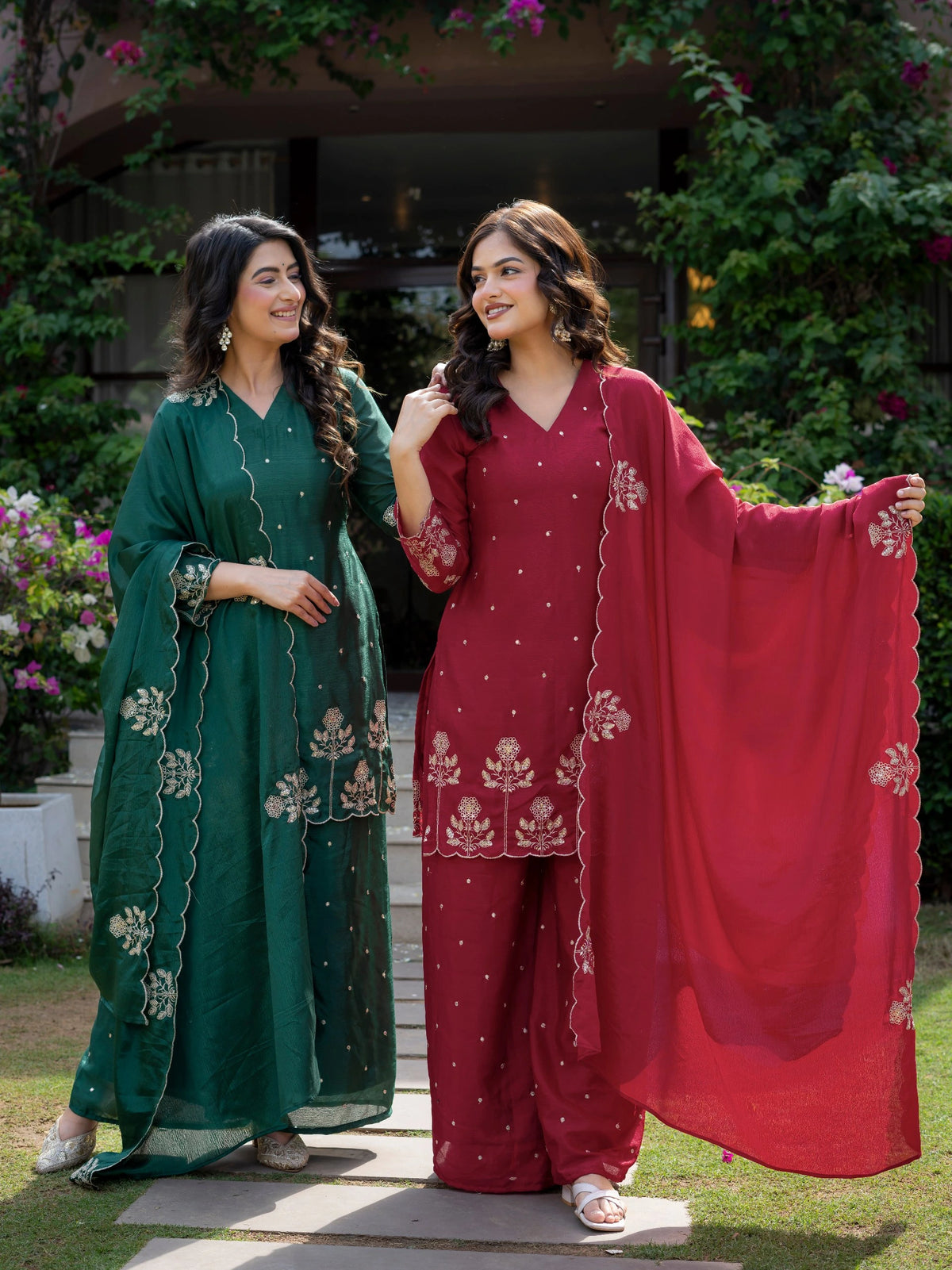 Two women in traditional outfits standing outdoors with greenery in the background