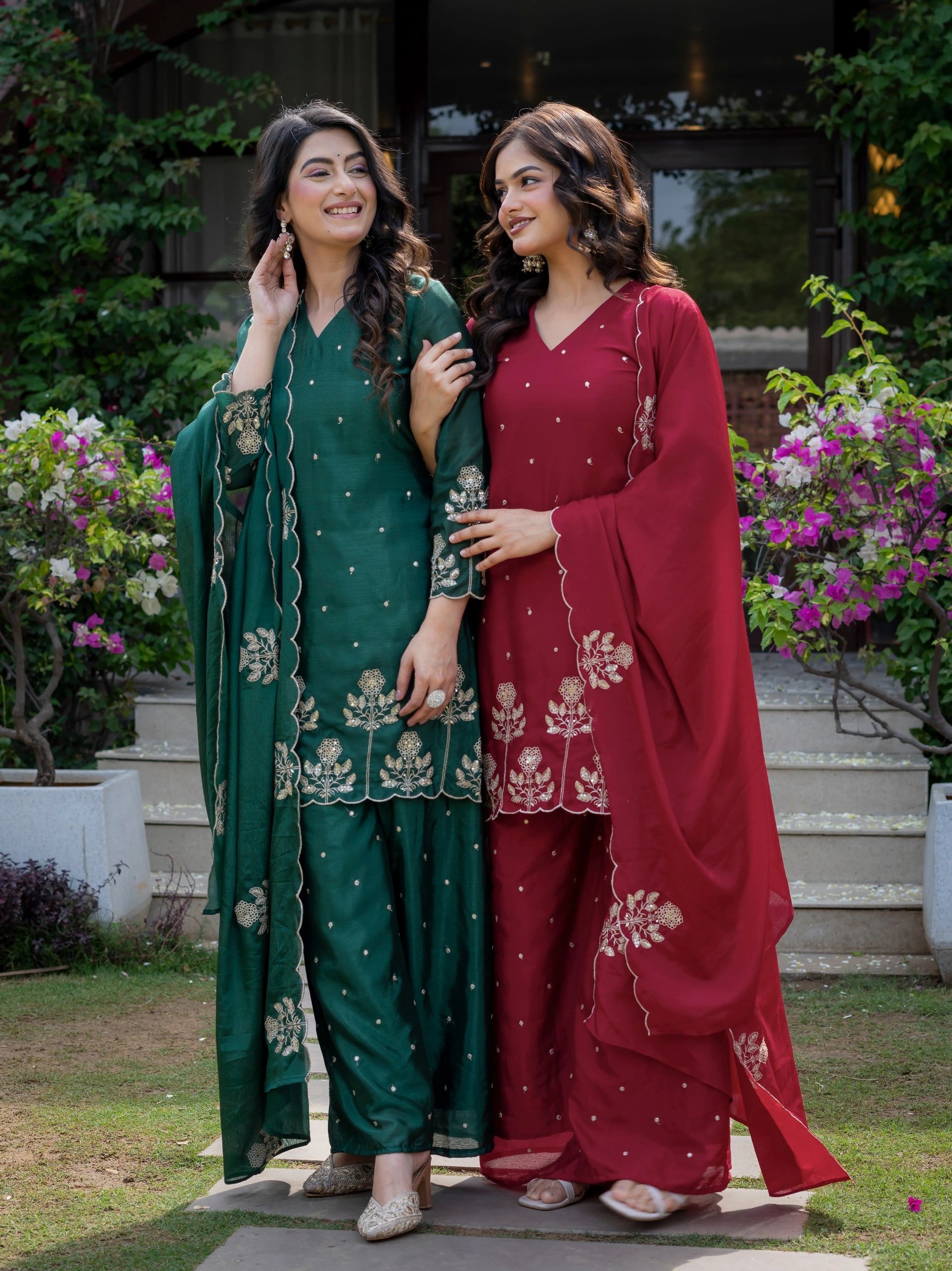 Two women in traditional green and red outfits standing outdoors with flowers and steps in the background.