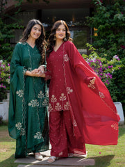 Two women in traditional green and red outfits standing outdoors with a garden background.
