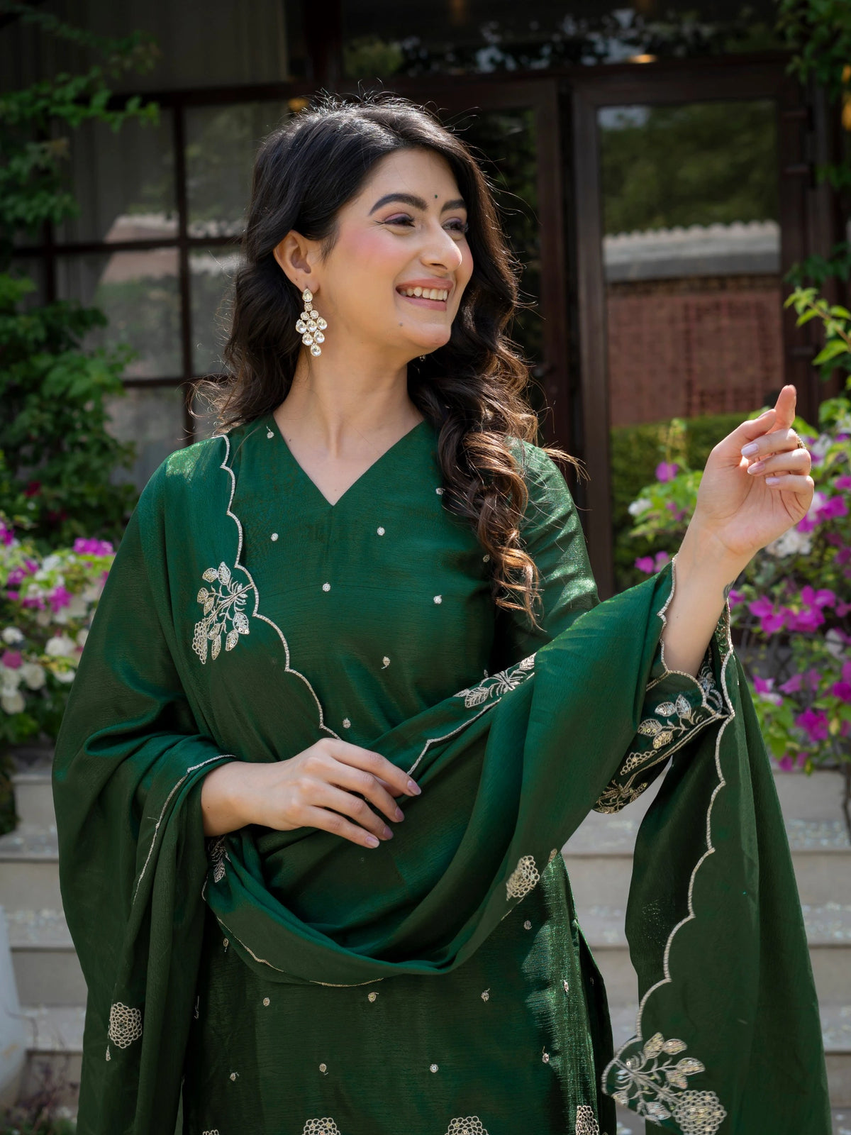 Woman wearing a green traditional outfit with floral patterns, standing outdoors with plants in the background.