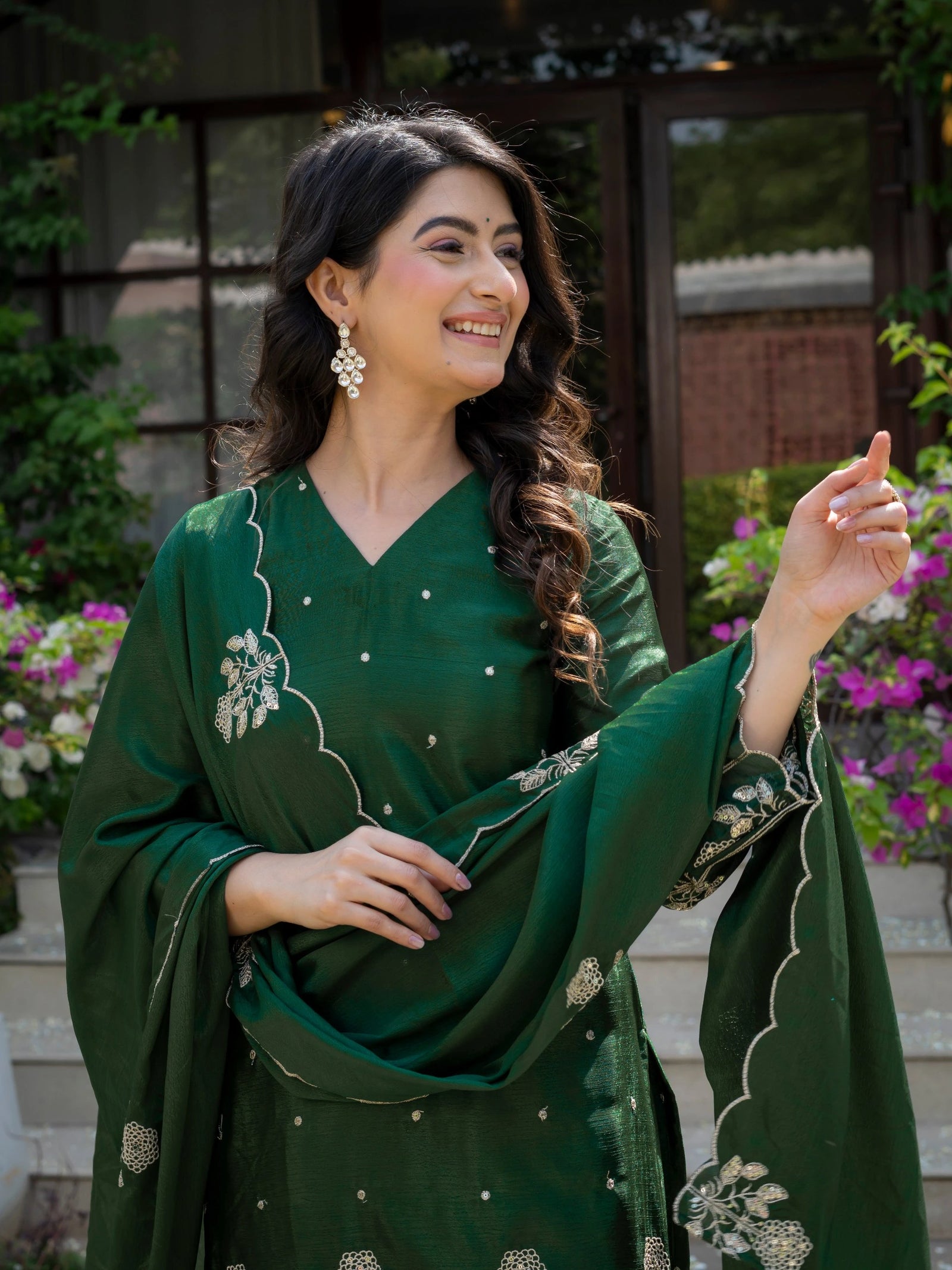 Woman wearing a green traditional outfit with floral patterns, standing outdoors with plants in the background.