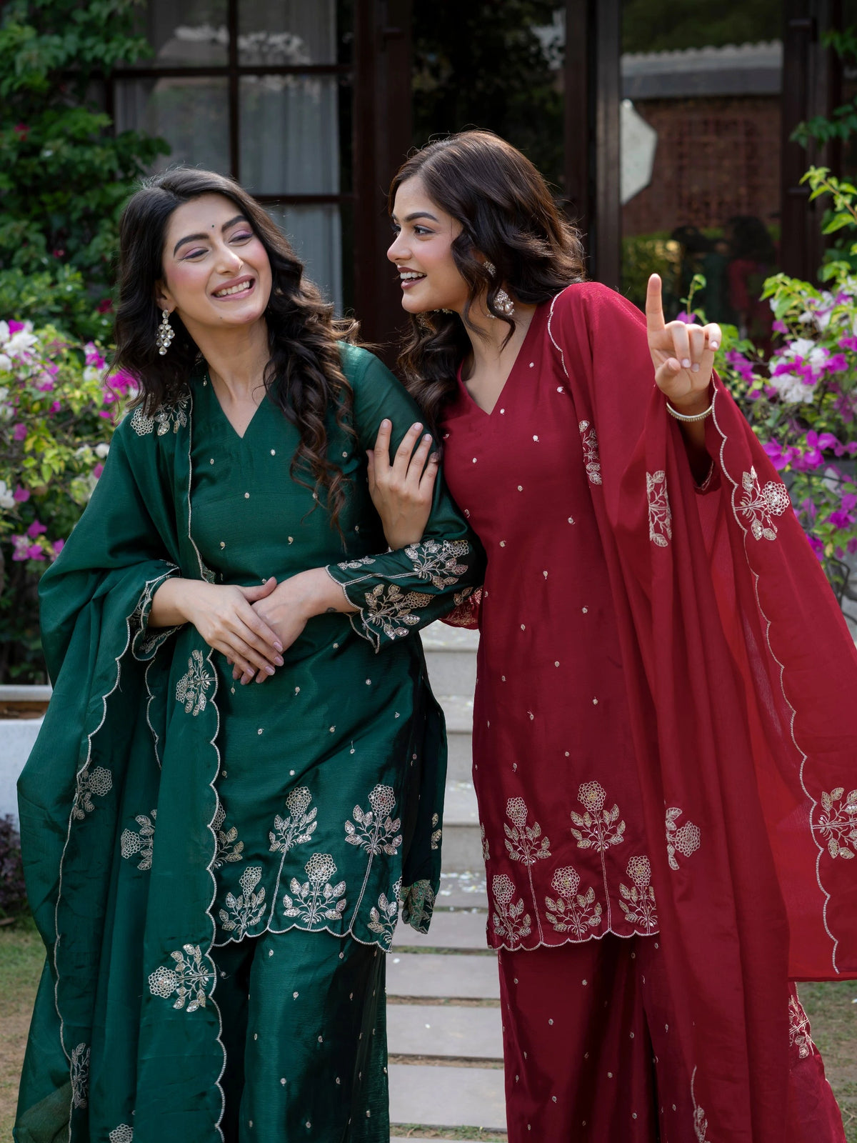 Two women in traditional green and red outfits standing outdoors with flowers in the background