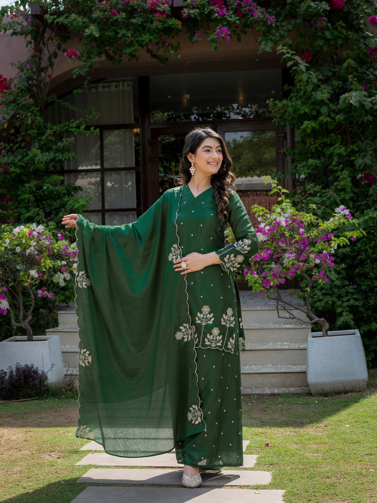 Woman in a green traditional outfit standing outdoors with flowers and plants in the background