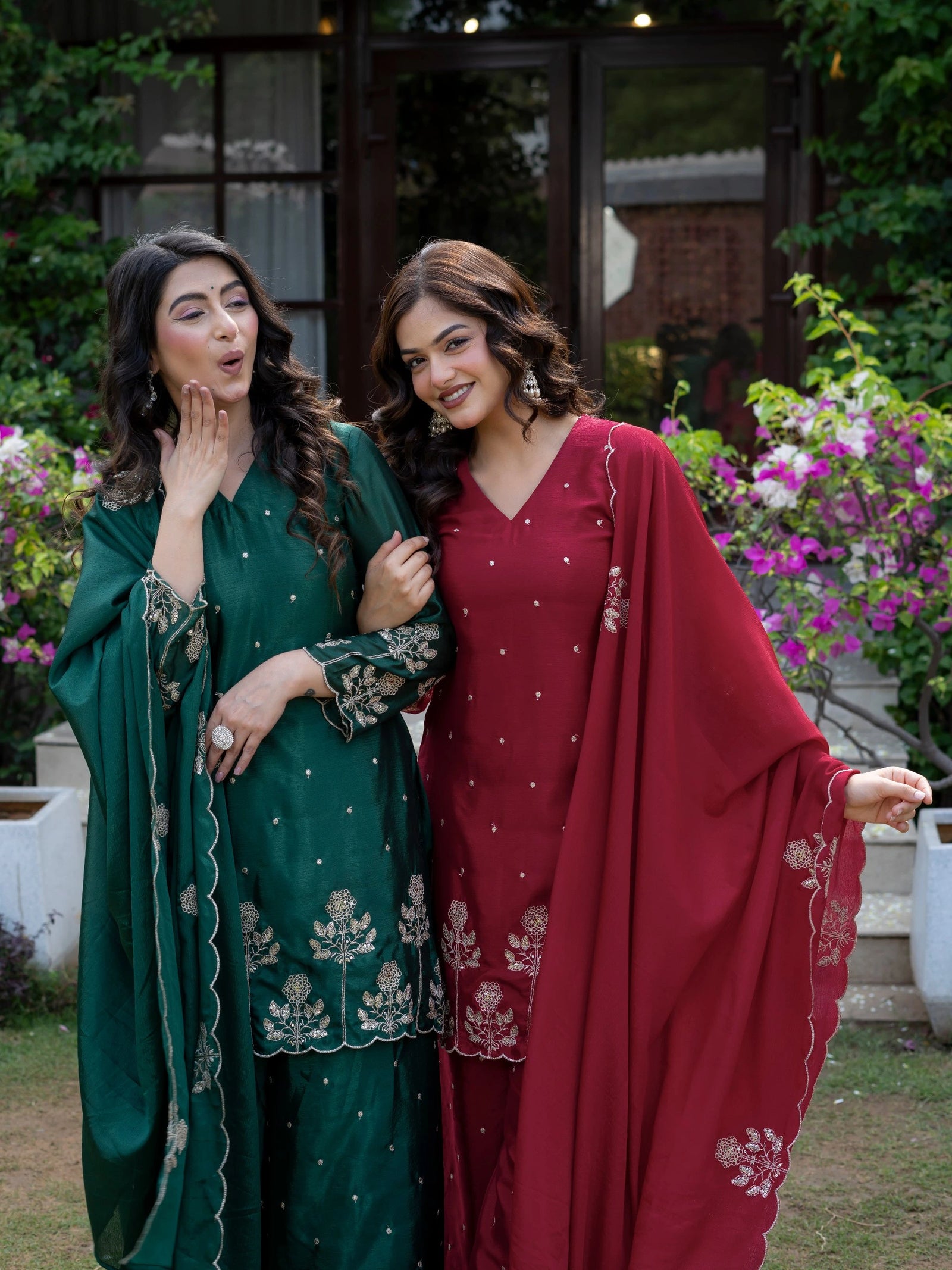 Two women in traditional green and red outfits standing outdoors with flowers and a building in the background.