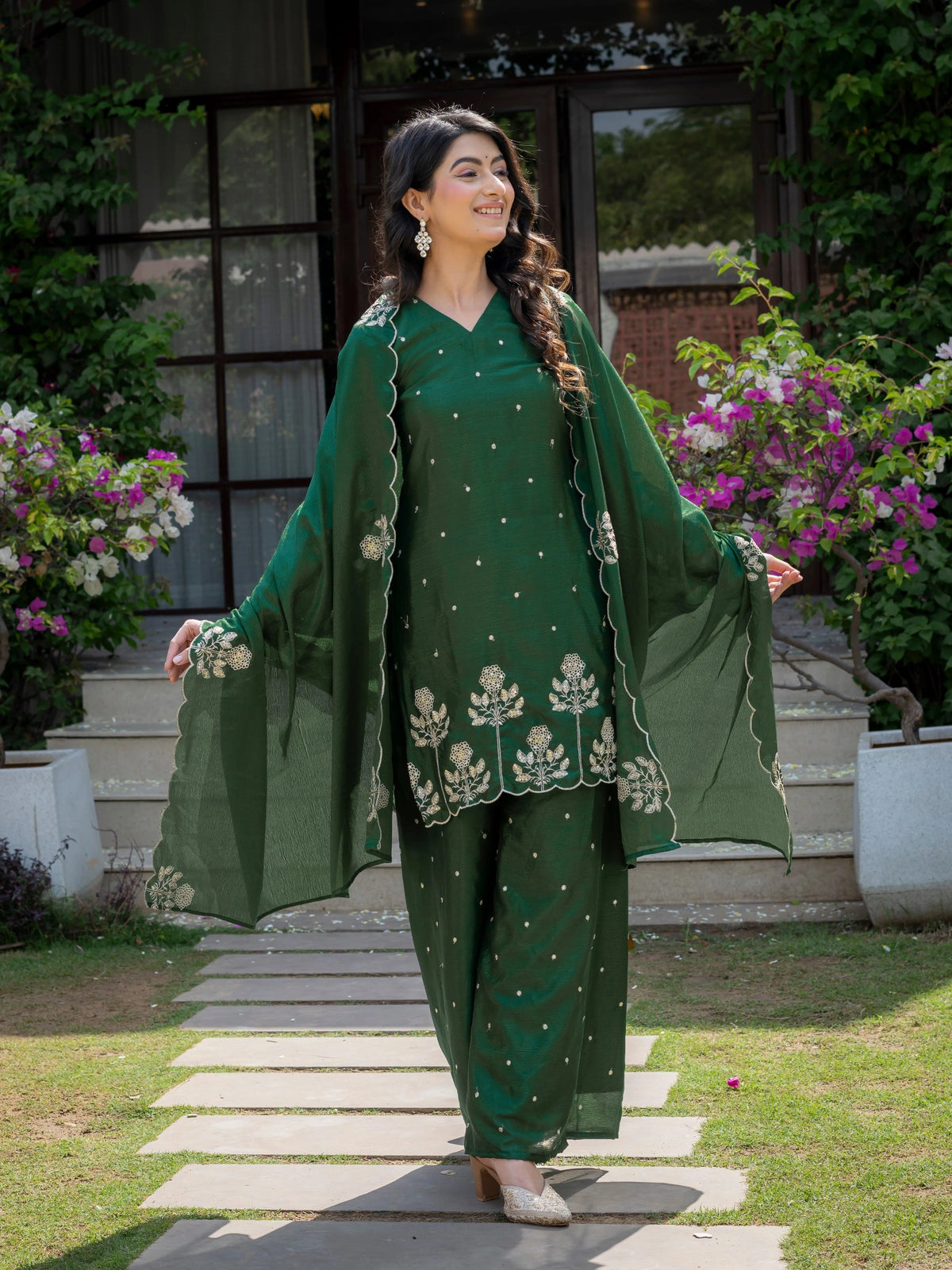 Woman in a green traditional outfit standing outdoors with plants and flowers in the background