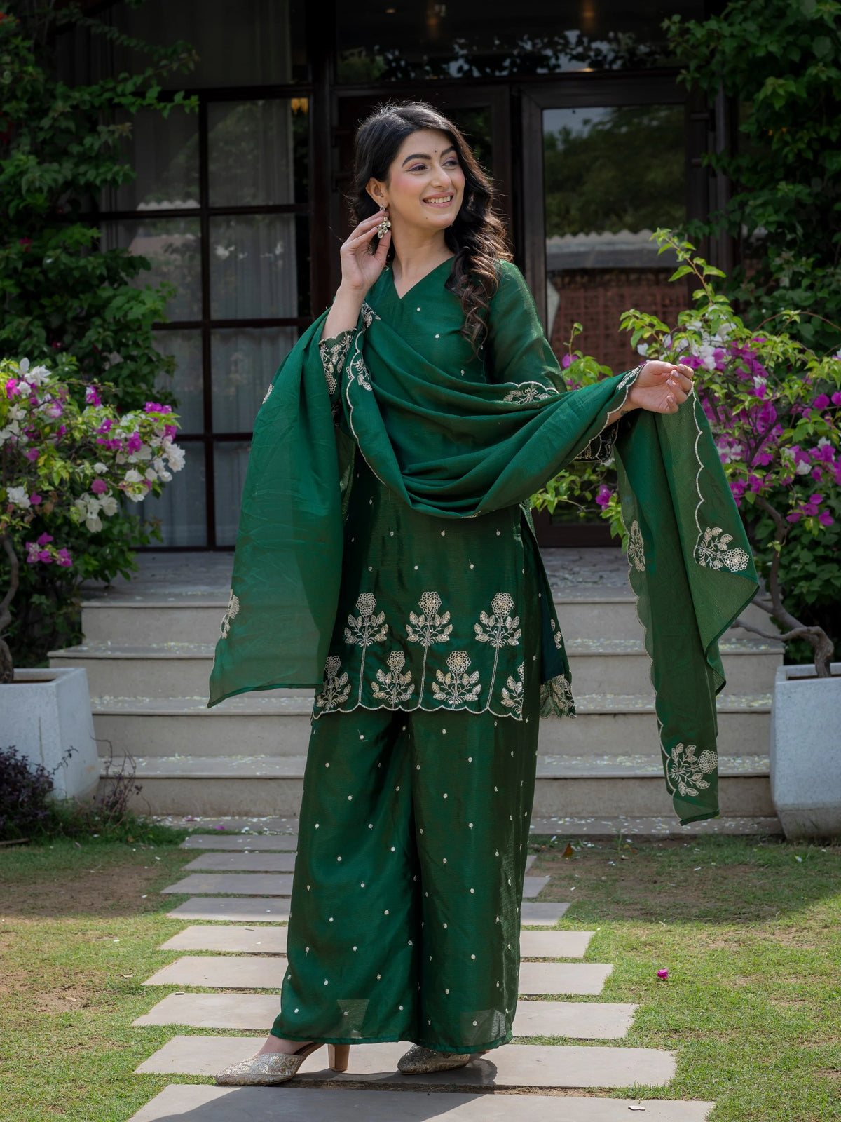 Woman in a green traditional outfit standing outdoors with flowers and steps in the background