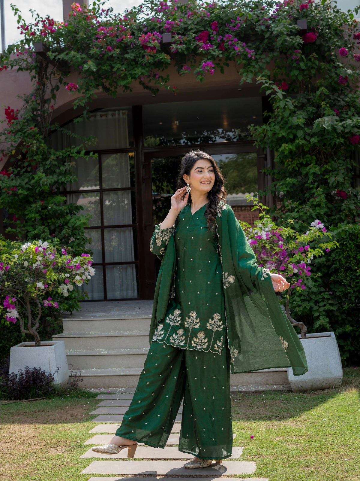 Woman in a green traditional outfit standing in a garden with flowers and a building in the background