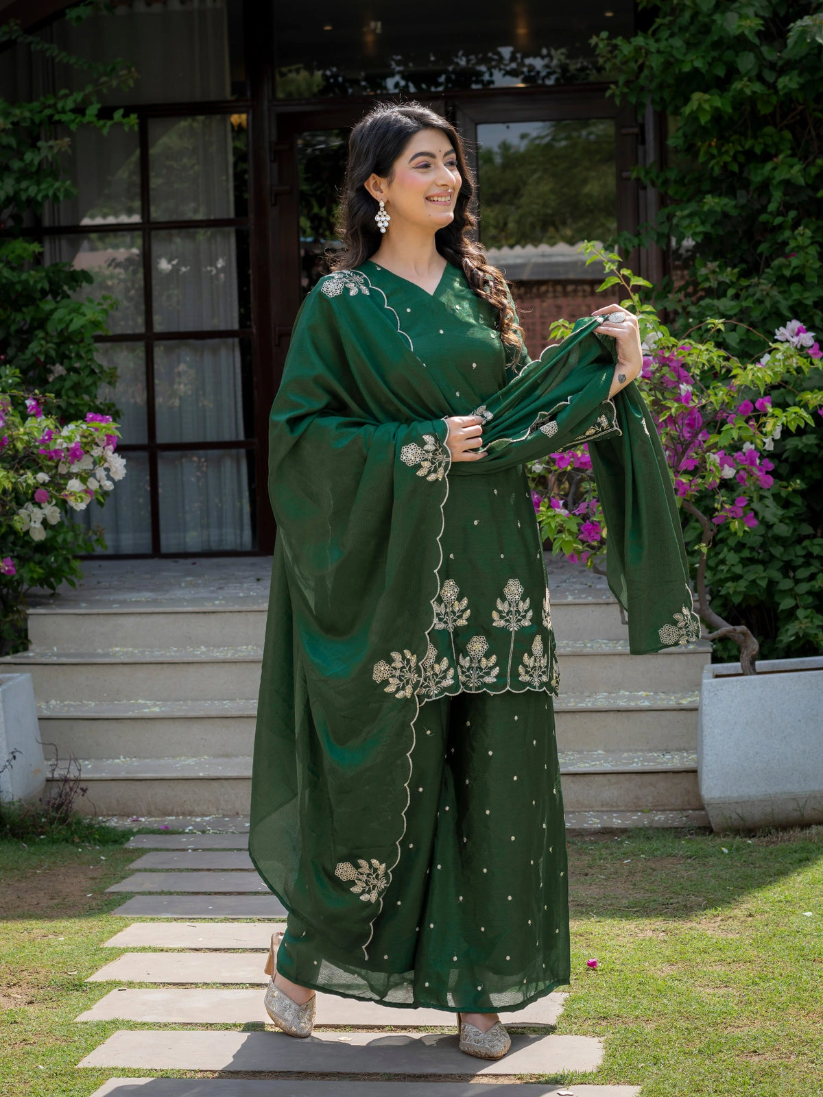 Woman in a green traditional outfit standing outdoors with flowers and a building in the background