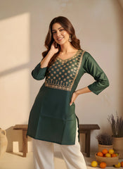 Woman wearing a green embroidered kurta with white pants in a home setting.