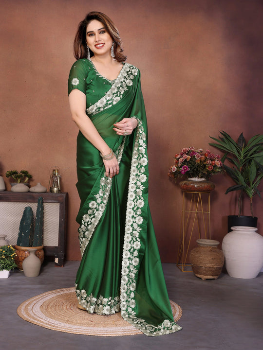 Woman in a green saree with floral patterns standing in a decorated room.