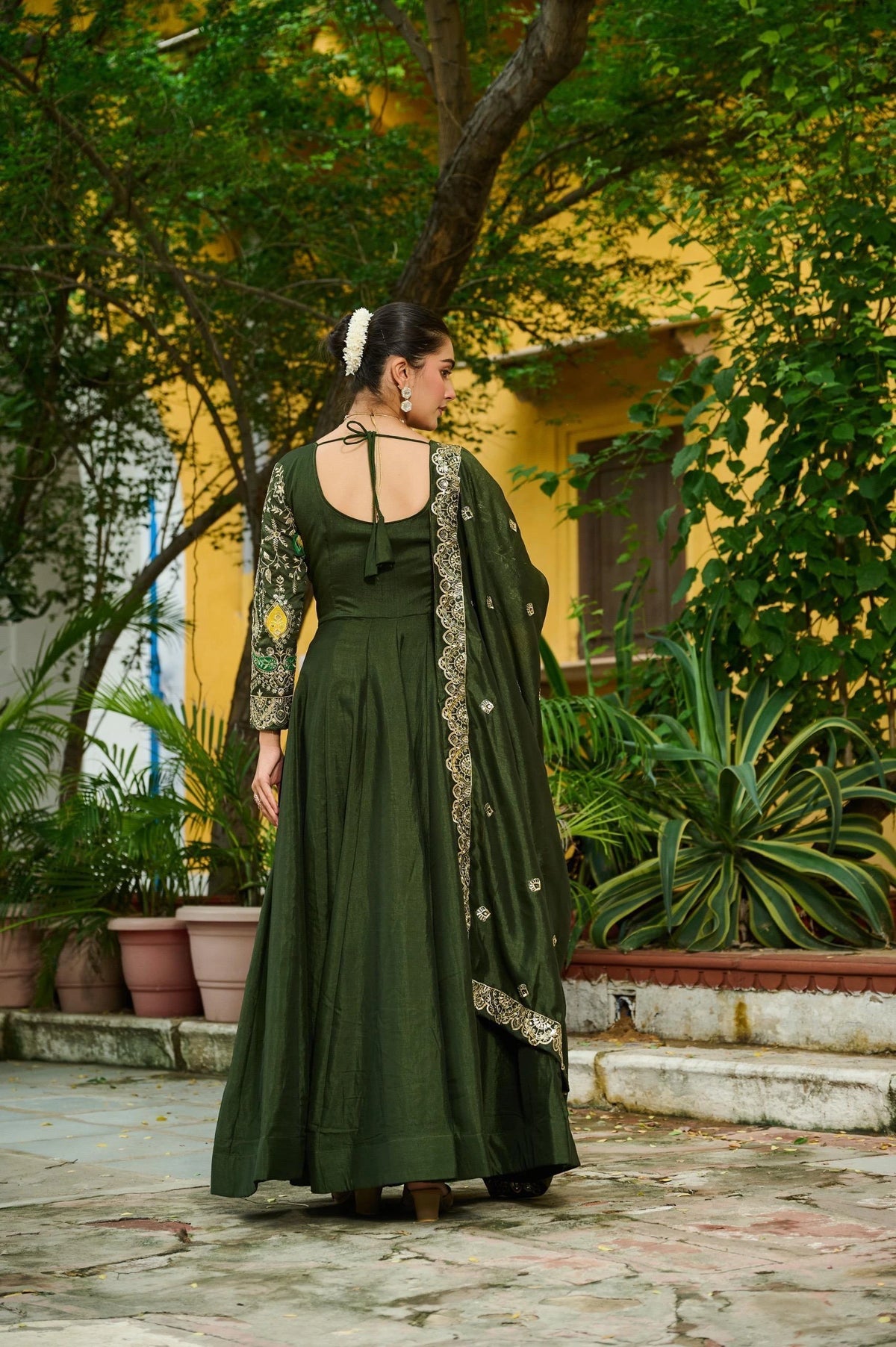 Woman in a green dress with floral patterns standing outdoors near a yellow building and plants.