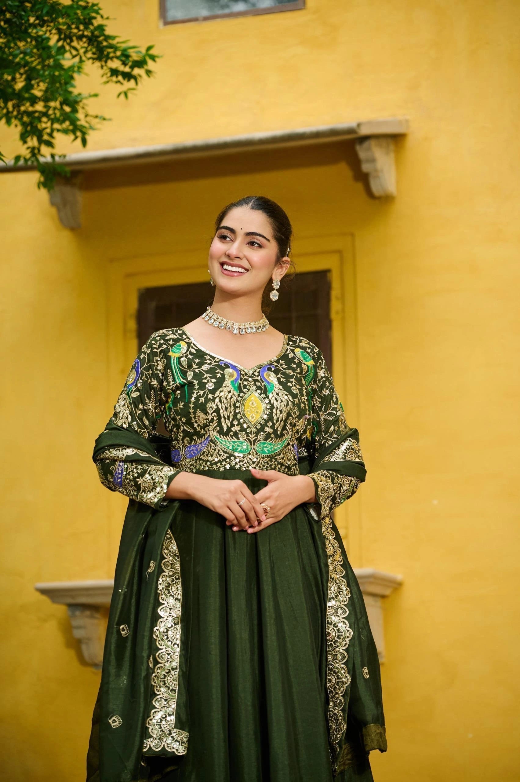 Woman in a green traditional outfit with intricate designs standing against a yellow wall.