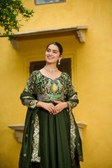 Woman in a green traditional outfit with intricate designs standing against a yellow wall.