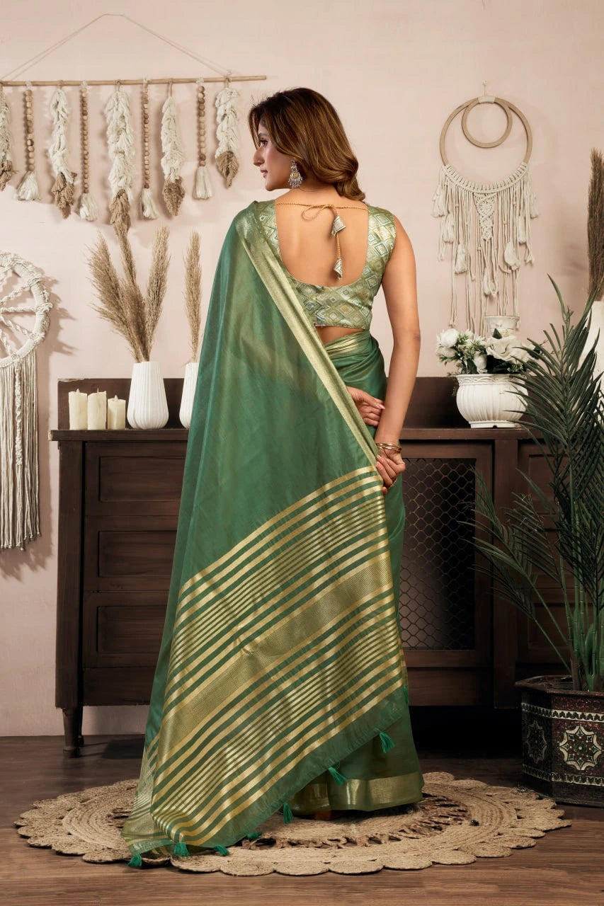 Woman in a green saree with gold patterns standing in a room with decorative elements.
